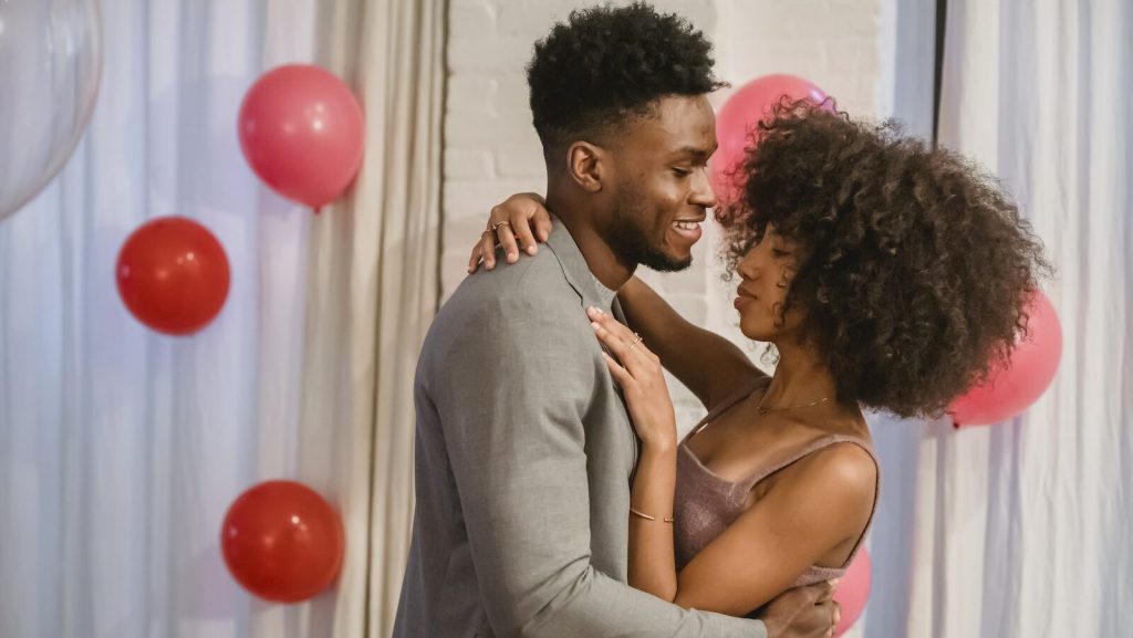 Side view of young black couple in elegant clothes standing in bright room and cuddling while dancing together with closed eyes near pink balloons on wall and curtains